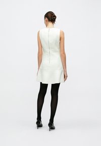 Biała, teksturowana sukienka bez rękawów z rozkloszowanym dołem, z centralnym zamkiem z tyłu, zestawiona z czarnymi, kryjącymi rajstopami i czarnymi szpilkami.