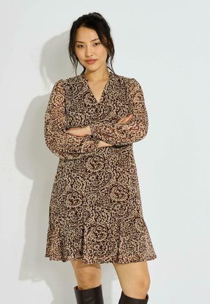 Mujer con cabello oscuro, vestido marrón con estampado y mangas largas, brazos cruzados, de pie frente a un fondo liso y claro, con botas negras.