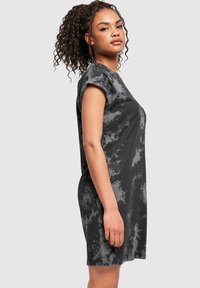 Kehaga naine, kes kannab lühikeste varrukatega musta ja halli lipsuvärvitud kleiti, seisab profiilis ühevärvilise tausta ees.