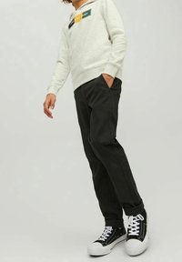 Person som bär en ljusgrå sweatshirt, svarta byxor med manschetter och svarta högskor med vita sulor och snörning, står med ena handen i fickan.