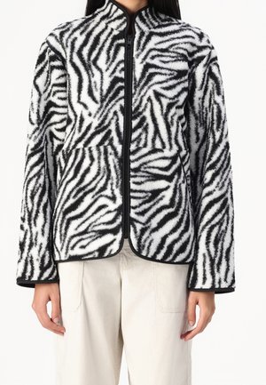 Kvinde iført en sort-hvid zebra-mønstret fleecetrøje med lynlås foran og beige bukser.