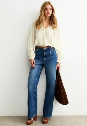 Blusa de crema clara con mangas abullonadas, vaqueros de pierna ancha azul, cinturón marrón, sandalias de plataforma y un bolso tote marrón. Tela texturizada en toda la prenda.