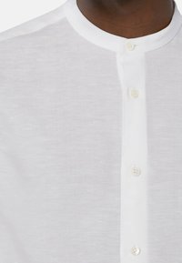 Camicia bianca in lino con colletto rotondo, dotata di una patta con bottoni e una texture sottile. I bottoni sono tondi e color crema.