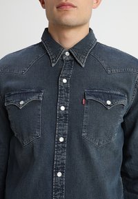 Homme portant une chemise en denim foncé à boutons, avec deux poches à rabat sur le devant, des boutons-pression blancs et une petite étiquette rouge sur la poche gauche.