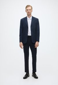 Fato navy com um blazer de um único botão, dois bolsos frontais e calças ajustadas a combinar, acompanhado de uma camisa branca de botão e sapatos pretos.