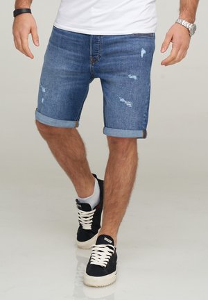 Shorts en denim bleu moyen avec des patches distressés, des ourlets roulés et un design classique à cinq poches, associés à des baskets noires.