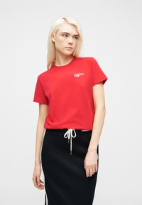 Ξανθιά γυναίκα που φοράει κόκκινο t-shirt Converse και μαύρη φούστα με λευκό κορδόνι, στέκεται μπροστά σε απλό λευκό φόντο.