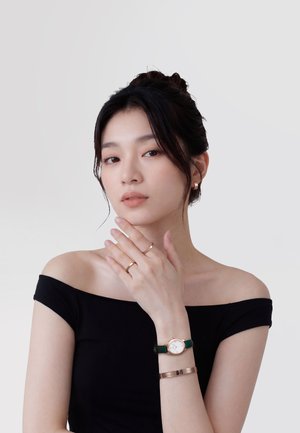 Tânără cu păr închis la culoare, purtând un top negru cu umerii goi, inele aurii, un ceas cu curea verde și o brățară din aur roz, pozând pe un fundal simplu.