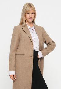 Cappotto beige a spina di pesce con vestibilità su misura, chiusura a un solo bottone e due tasche laterali. Indossato sopra una camicia lilla e una gonna nera.
