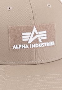 Căciulă de baseball khaki cu un patch texturat din fetru bej, având un logo alb embosat și numele brandului "ALPHA INDUSTRIES." Design structurat.