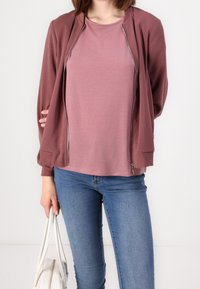 Burgundy bomber jacket with zip closure and ribbed cuffs, layered over a light pink t-shirt, paired with blue jeans and a white handbag.