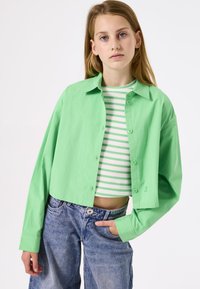 Mintgrünes kurzes Hemd mit Knopfleiste und langen Ärmeln über einem weißen und grünen gestreiften Cropped-Top, kombiniert mit hoch taillierten blauen Jeans.