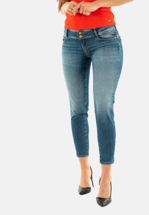 Jeans de denim azul delgados con un acabado desgastado, dos bolsillos frontales, herrajes dorados y una cintura de altura media, llevados con tacones altos negros.