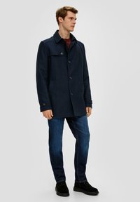 Marineblauwe jas met een dubbelgebreide ontwerp, knoopsluitingen en zijzakken; gecombineerd met blauwe jeans en zwarte schoenen. Gladde, gestructureerde stof.