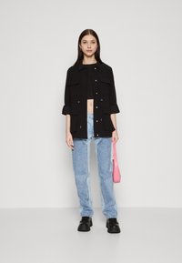 Giacca camicia nera con bottoni, dotata di due tasche frontali, indossata sopra un top corto, abbinata a jeans larghi azzurri e scarpe chunky nere.