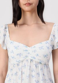 Abito bianco con motivo floreale blu, maniche a palloncino corte, scollo quadrato, tessuto stropicciato e dettagli arricciati sul seno.