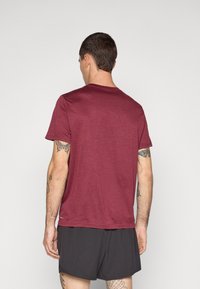 Bordeauxfarbenes kurzärmliges T-Shirt aus weichem Stoff, mit Rundhalsausschnitt und lockerer Passform, kombiniert mit dunklen Shorts und sichtbaren Tätowierungen an den Armen.