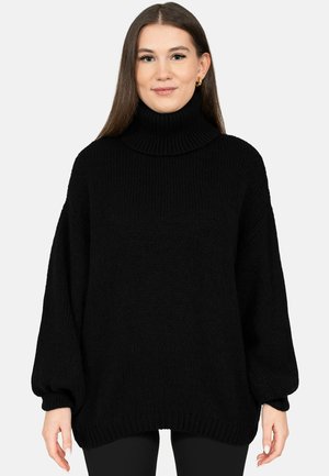 Femme aux longs cheveux bruns portant un pull noir en grosse maille à col roulé avec des manches ballon sur un fond clair uni.