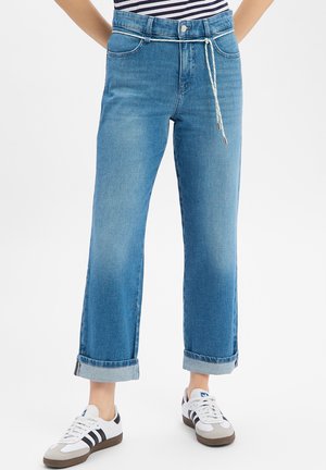 Femme portant un jean bleu retroussé avec une ceinture à cordon et des baskets blanches avec des rayures noires, debout sur un fond blanc.