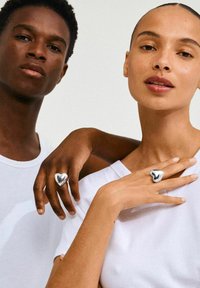 To personer iført hvide t-shirts holder hænder og viser sølvringe med hjerteform og et flydende design. Ringene er glatte og reflekterende.