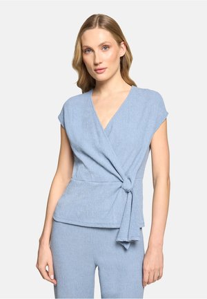 Vrouw die een lichtblauw getextureerd overslagtopje met korte mouwen en bijpassende broek draagt, staande tegen een effen lichte achtergrond.