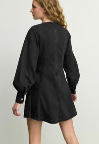 Mustapillinen denim-mekko, jossa on pitkät puffihihat, pyöreä pääntie ja vetoketju takana. Mekko esittelee ommeltuja sauma- ja napillisia hihansuita.