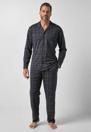 Homme barbu portant un pyjama à carreaux bleu foncé et marron à manches longues boutonné avec un pantalon assorti, debout pieds nus sur un fond clair.