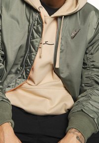 Giacca bomber in nylon verde oliva con chiusura frontale con zip, polsini a coste e fodera arancione, indossata sopra una felpa con cappuccio beige con logo nero.