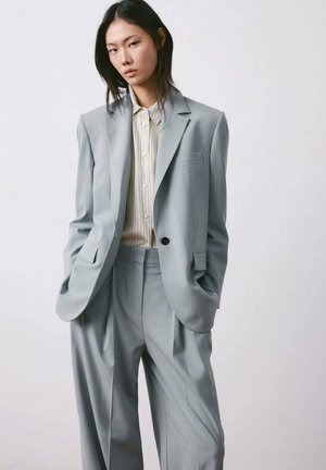 Joven mujer de pie con las manos en los bolsillos, vistiendo un traje gris claro de gran tamaño y una camisa de botones a rayas, contra un fondo liso.