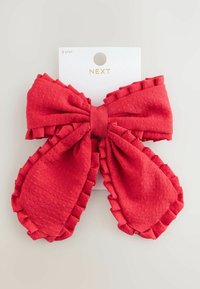 Noeud de cheveux rouge en tissu texturé avec des bords froncés, suspendu à une carte blanche étiquetée "NEXT" avec "3 ans+" en haut.