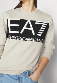 Beige långärmad sweatshirt med en svart rektangulär panel med vit text "EA7" och "Emporio Armani", rund halsringning och ribbad mudd.