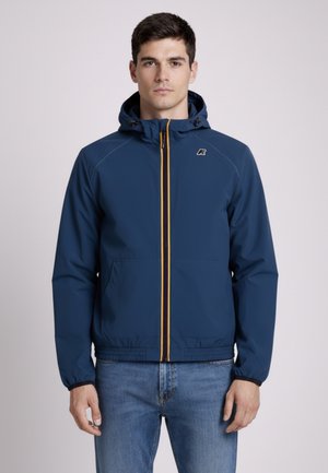 Uomo che indossa una giacca con cappuccio blu navy con cerniera arancione e jeans blu, in piedi contro uno sfondo chiaro e uniforme.