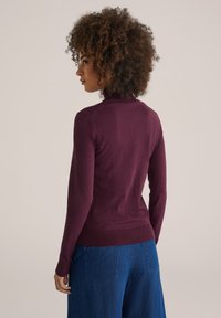 Bordeauxfarbener, eng anliegender Pullover mit langen Ärmeln und hohem Kragen, kombiniert mit weiten blauen Jeanshosen, die eine glatte Textur aufweisen.