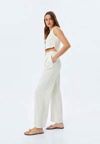 Witte linnen cropped top met golvende zoom, hoge linnen broeken en sandalen met bandjes en een decoratieve bloemdetail.