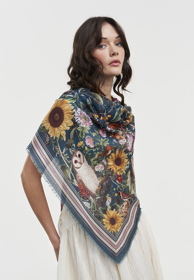 Woman with wavy brown hair wears a dark blue floral scarf featuring sunflowers, birds, and an owl over a white dress.