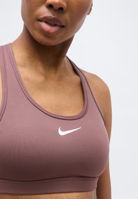 Nærbillede af en person iført en mauve Nike sports-bh med en hvid swoosh-logo på en ensfarvet baggrund.