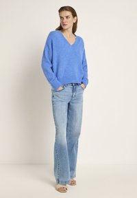 Hellblaues Strickpullover mit V-Ausschnitt, kombiniert mit hellblauen Schlagjeans. Der Pullover hat gerippte Bündchen und einen gerippten Saum. Keine Muster oder sichtbare Hardware.