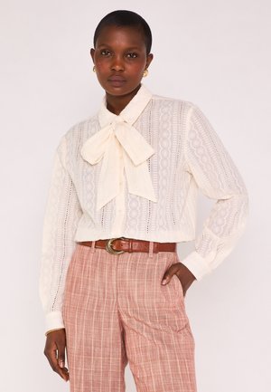 Femme portant un chemisier transparent crème à motifs avec un grand nœud papillon, un pantalon rayé rose taille haute, une ceinture marron et des boucles d'oreilles dorées, debout la main dans la poche.