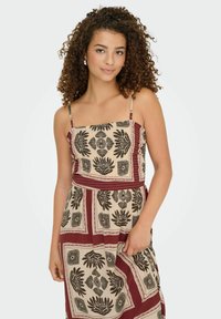 Femme jeune avec des cheveux bouclés portant une robe sans manches beige et bordeaux à motifs, posant avec une main tenant la jupe.