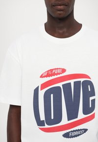 Λευκό βαμβακερό t-shirt με το εντυπωσιακό γράφημα "LOVE" σε ναυτικό και κόκκινο χρώμα, με την ετικέτα "100% PURE" από πάνω και την ένδειξη "Fiorucci" από κάτω σε κυκλικό σχέδιο.