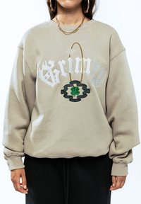 Beige sweatshirt met een oversized pasvorm, voorzien van witte "Grimy" tekst en een groene geborduurde klavertje-detail, aangevuld met een gouden ketting accent.