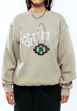 Sudadera beige de corte oversize, con texto blanco "Grimy" y un detalle de trébol bordado en verde, complementada por un acento de cadena dorada.