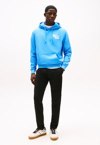 Jauns vīrietis stāv valkādams spilgti zilu Tommy Jeans kapuci, melnas bikses un baltas sporta kurpes ar gumijas zolēm uz baltas fona.