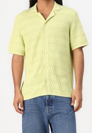 Mann trägt ein hellgrünes kurzärmeliges Strickhemd mit Knopfleiste und strukturiertem Muster und blaue Jeans, steht vor einem schlichten weißen Hintergrund.