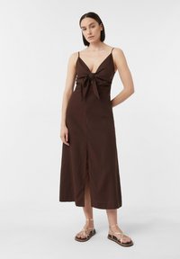 Braunes Midi-Kleid mit tiefem V-Ausschnitt und vorderer Schleifendetail. Dünne Träger, gerade Silhouette und ein vorderer Schlitz. Kombiniert mit goldenen Sandalen.