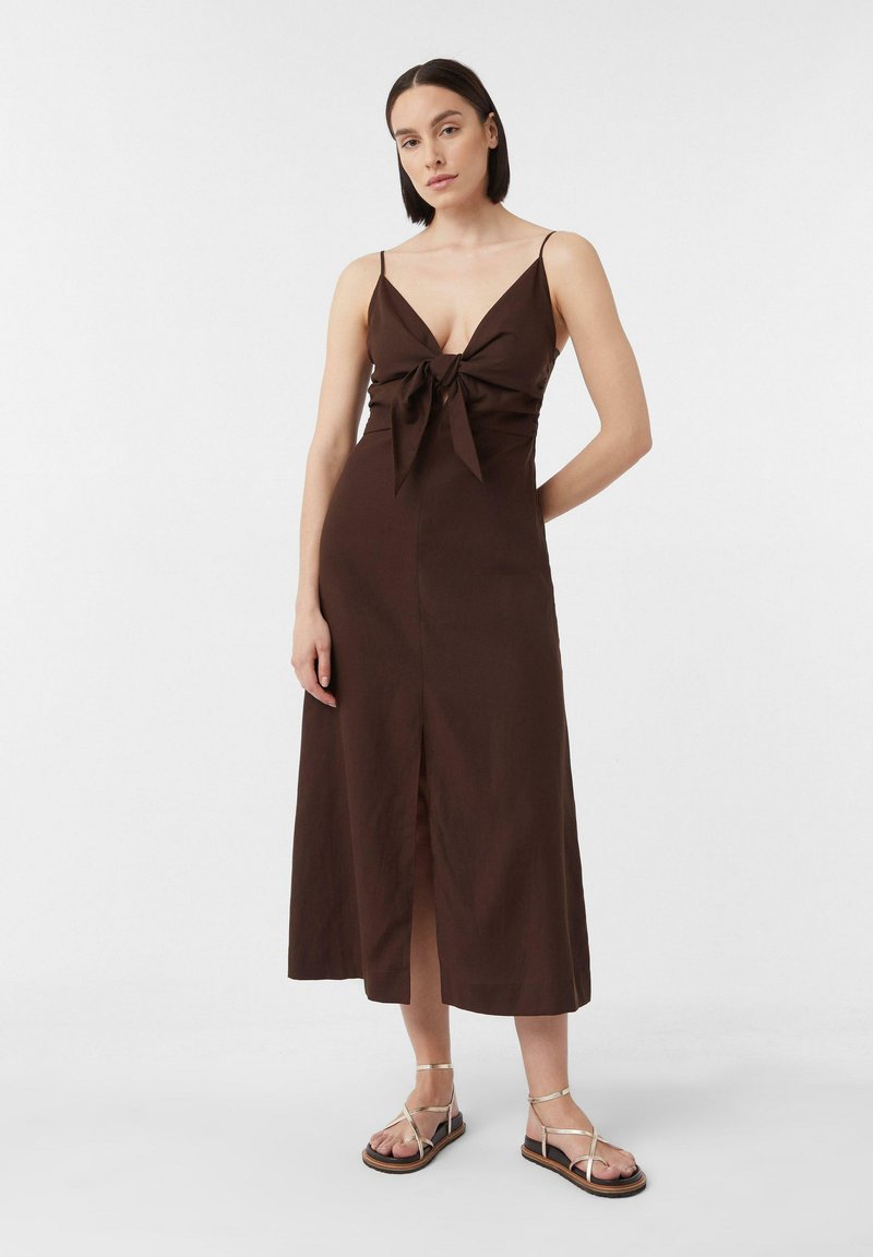 Braunes Midi-Kleid mit tiefem V-Ausschnitt und vorderer Schleifendetail. Dünne Träger, gerade Silhouette und ein vorderer Schlitz. Kombiniert mit goldenen Sandalen.