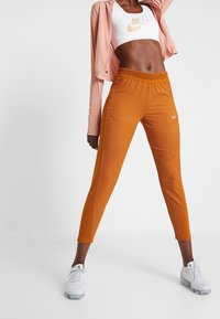 Orange Sporthosen mit glatter Textur, seitlichen Mesh-Akzenten und kleinem weißen Nike-Logo. Kombiniert mit einer rosa Cropped-Jacke und weißen Schuhen.
