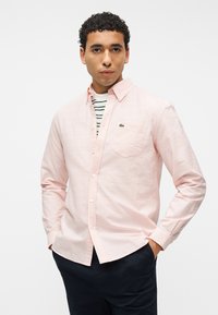 Jeune homme aux cheveux bouclés portant une chemise rose clair à boutons par-dessus un t-shirt rayé blanc et vert, les mains dans les poches d'un pantalon foncé.