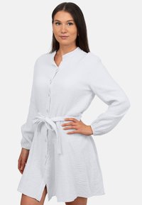 Abito bianco a camicia realizzato in tessuto testurizzato, con colletto cinese, maniche lunghe e una cintura annodata in vita.