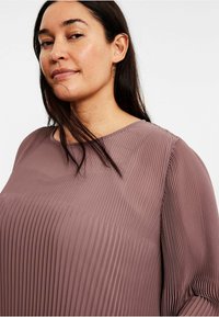 Plisséblus i mauve tyg, med vertikala veck, bred halsringning och långa ärmar. Slät textur med en avslappnad passform.